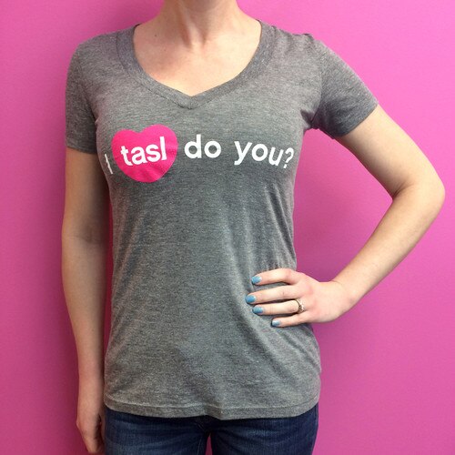 An image of a woman wearing the dark gray fitted tasl v-neck t-shirt standing against a pink background.