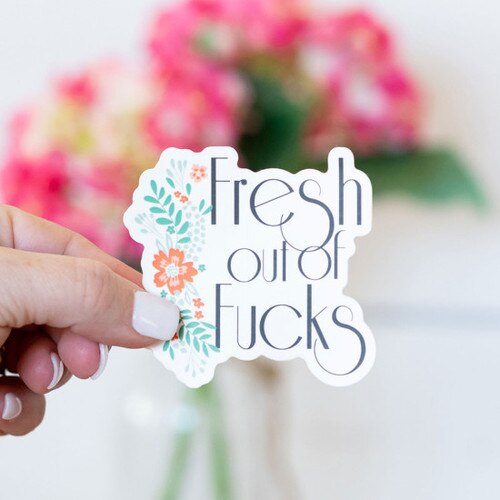 Focused on someone holding a Fresh Out Of Fucks sticker with flowers in the background