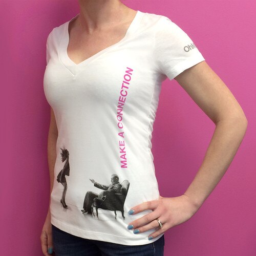 An image of the torso of a woman wearing the white V-neck Make a Connection OhMiBod t-shirt standing against a pink background.