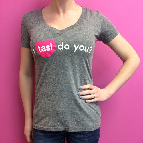 An image of a woman wearing the dark gray fitted tasl v-neck t-shirt standing against a pink background.